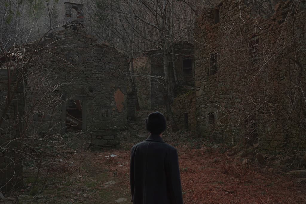 Figure seen from behind facing ruined stone buildings in a leaf-covered clearing.